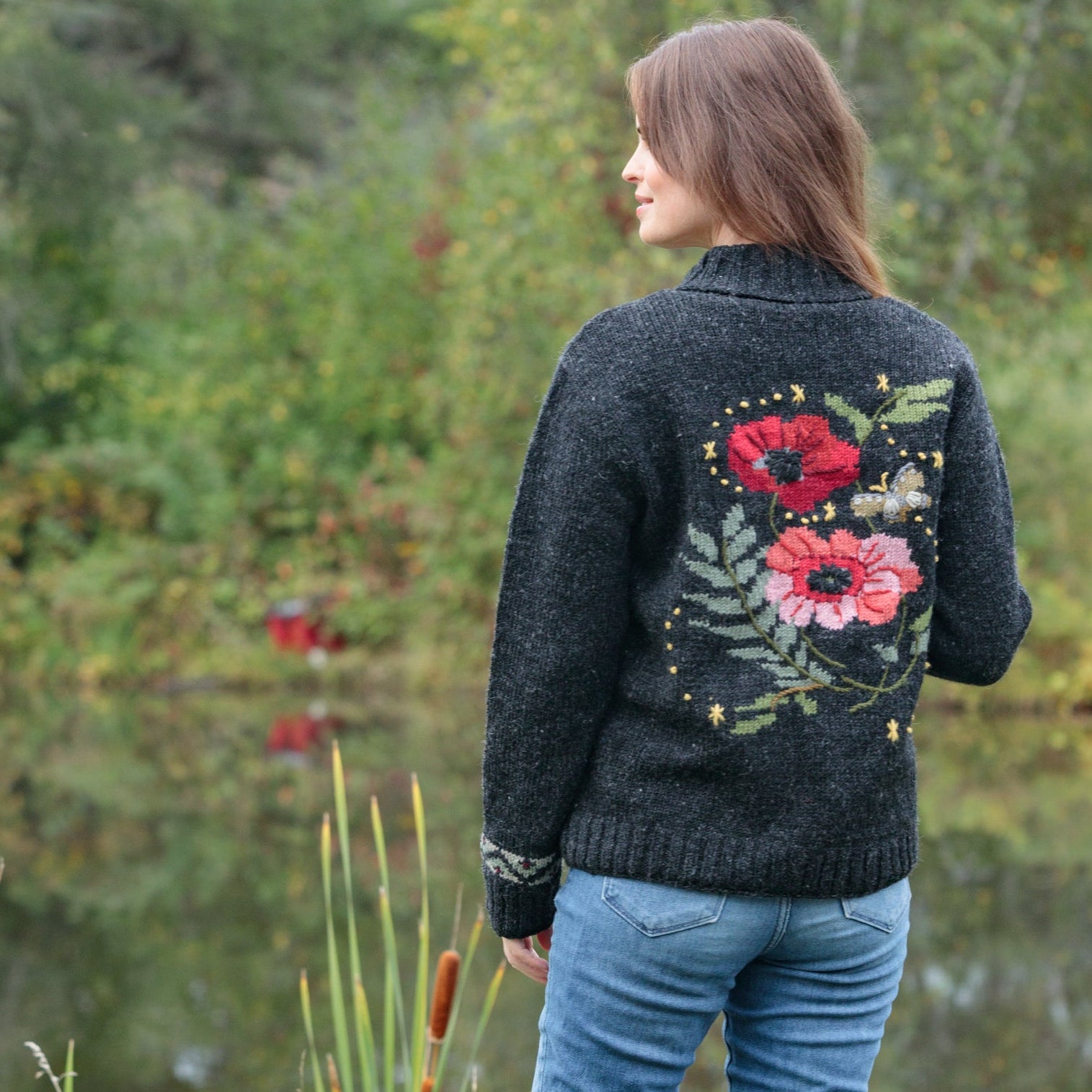 A woman standing outdoors by a pond, viewed from the back, wearing a dark charcoal knit sweater with a large red poppy and green leaf floral design on the back, paired with blue jeans and black boots.