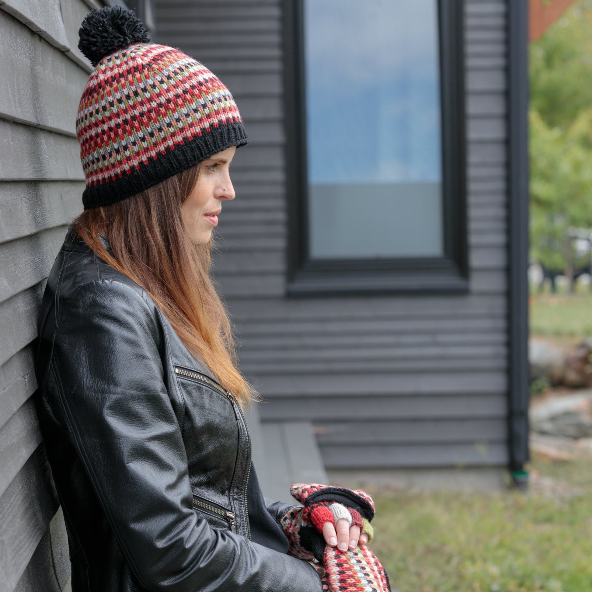 Person wearing a patterned beanie and leather jacket leaning against a building exterior.