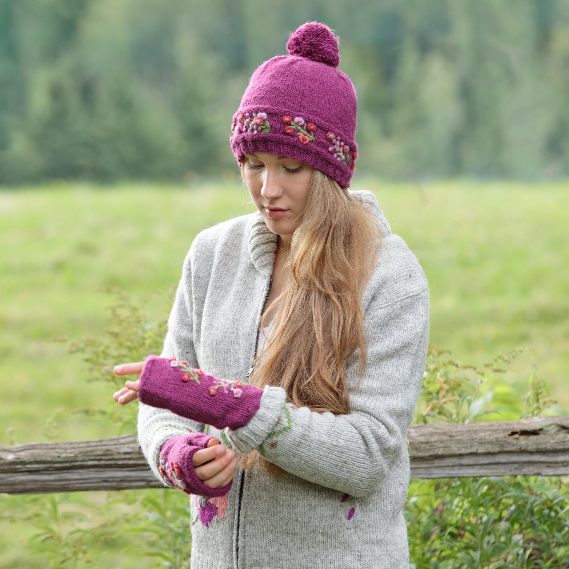 A woman standing outdoors in front of a wooden fence and green field, wearing a bright pink knit beanie and handwarmers with floral embroidery trim.