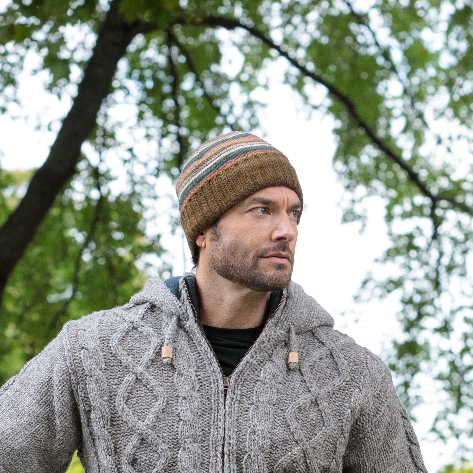 Man wearing a striped beanie and cable knit sweater outdoors with trees in the background