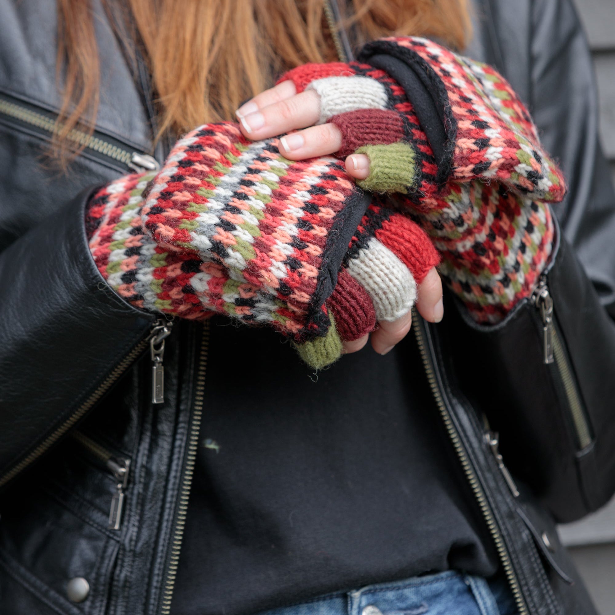 A close-up of a person wearing a black leather jacket, black top, and blue jeans, holding a pair of multicolor knit fingerless gloves with red, green, beige, and black geometric patterns.