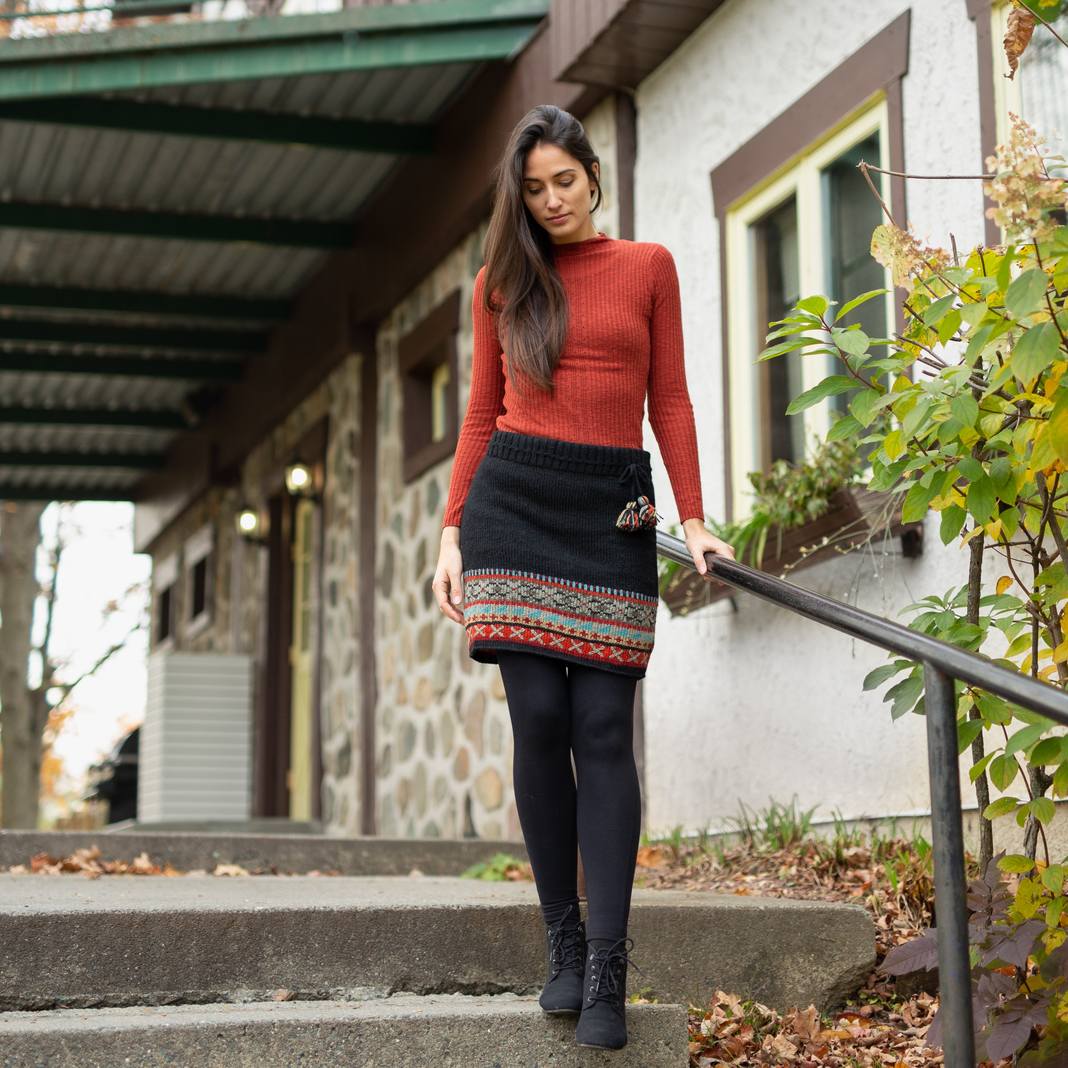 A black wool skirt with a Fair Isle pattern featuring red, blue, and white geometric designs, and a hand-stitched cross detail at the waist. An adjustable drawstring cord with a wool tassel is also visible.